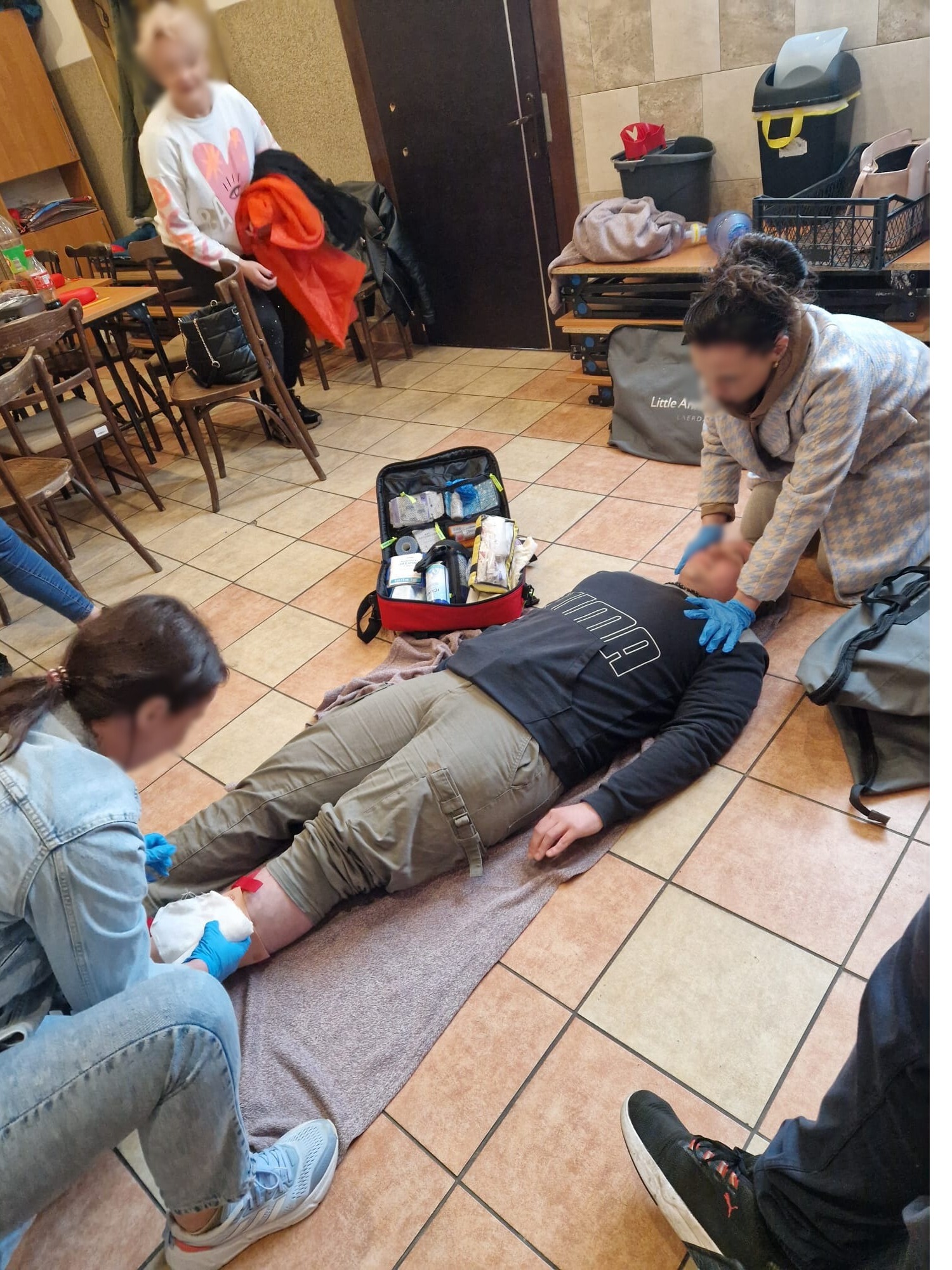 Osoby w trakcie udzielania pierwszej pomocy poszkodowanemu z zakrwawioną nogą. Widoczna apteczka, rękawice ochronne i opatrunki. Szkolenie z zakresu ratownictwa.