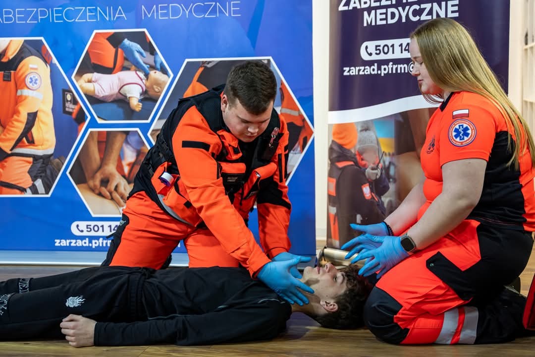 Instruktorzy w pomarańczowych uniformach demonstrują resuscytację krążeniowo-oddechową na manekinie podczas szkolenia z pierwszej pomocy medycznej.