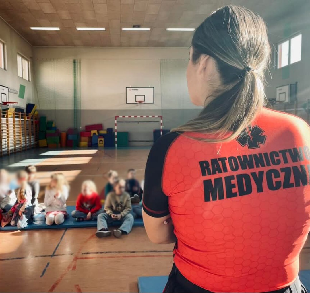 Instruktorka w czerwonej koszulce z napisem 'Ratownictwo Medyczne' prowadzi zajęcia z pierwszej pomocy dla grupy dzieci siedzących na podłodze sali gimnastycznej.