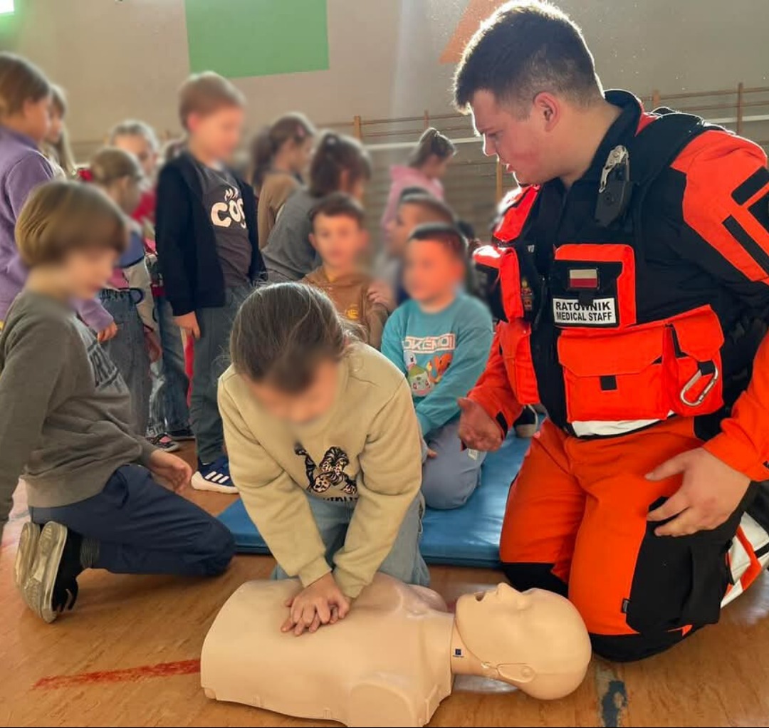 Ratownik w stroju uczy dzieci resuscytacji na manekinie podczas kursu. Dziecko wykonuje uciśnięcia klatki piersiowej, a w tle grupa obserwuje.