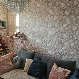 Stanisław Kościański - Salon z wzorzystą tapetą w kwiaty, szarą sofą z dekoracyjnymi poduszkami, choinką i ozdobnym żyrandolem.