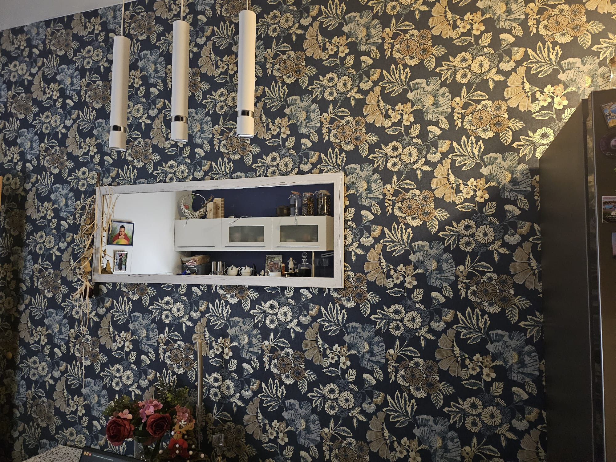 Ściana po remoncie z nową tapetą w kwiaty, oświetleniem i dekoracyjną ramą eksponującą zawartość szafek kuchennych.