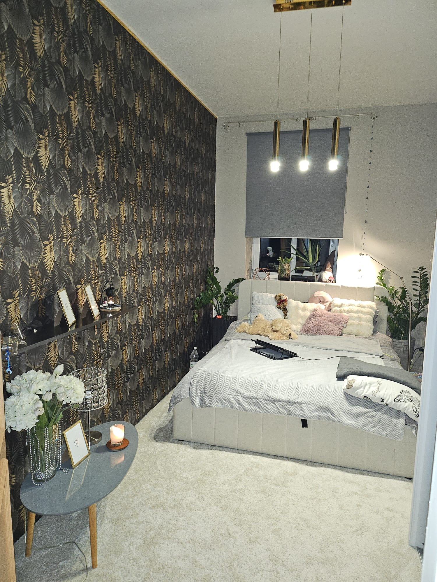 Wykończona sypialnia z tapetą w liście, łóżkiem z szarym nakryciem, roletą w oknie, stolikiem nocnym z kwiatami i oświetleniem.