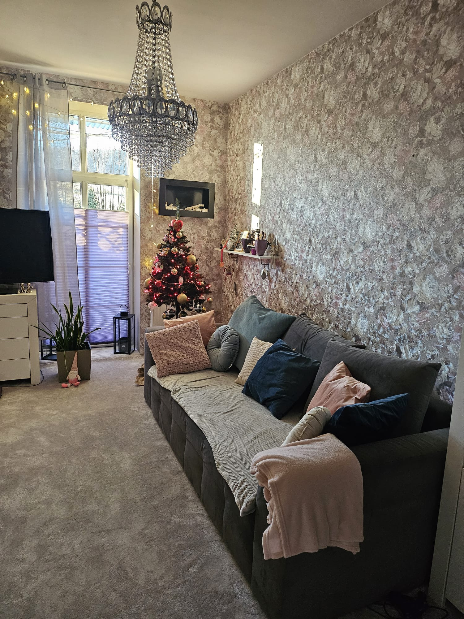 Salon w okresie świątecznym: ciemnoszara sofa z poduszkami i kocem, tapeta w kwiaty, kryształowy żyrandol, choinka z czerwonymi światełkami i ozdobami, biała komoda z telewizorem i rolety w oknie.