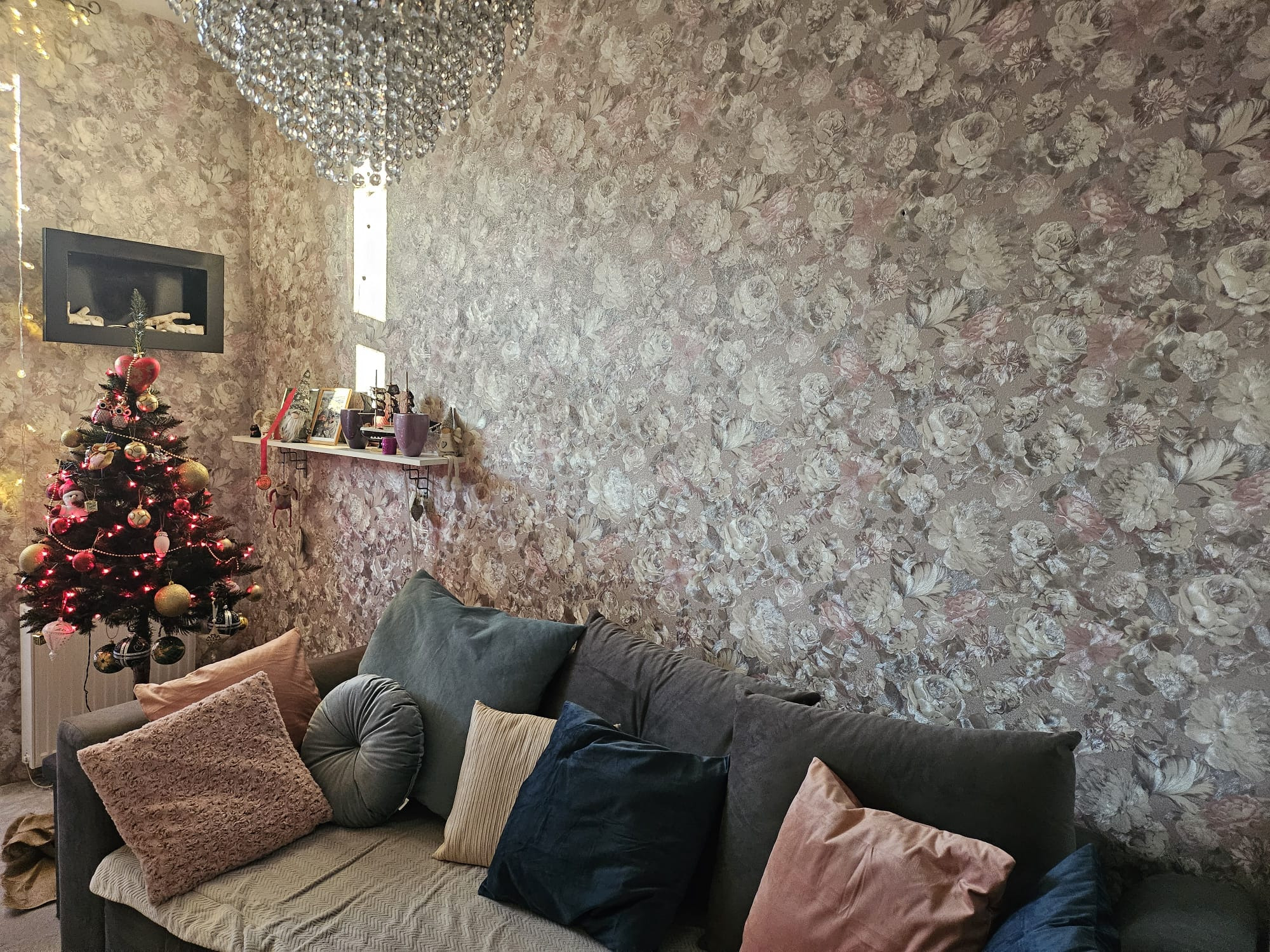 Salon z wzorzystą tapetą w kwiaty, szarą sofą z dekoracyjnymi poduszkami, choinką i ozdobnym żyrandolem.