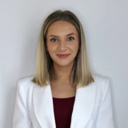 Portret uśmiechniętej blondynki w białym żakiecie i bordowej bluzce na jasnym tle. Profesjonalny wizerunek agenta nieruchomości, budzący zaufanie.