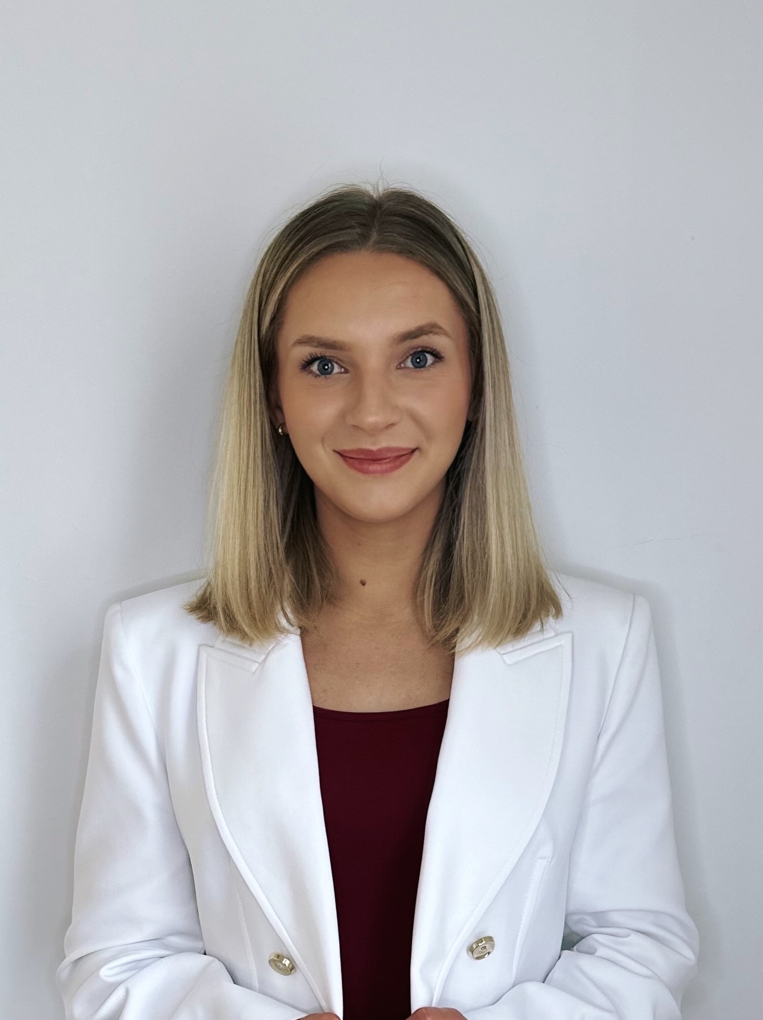 Portret uśmiechniętej blondynki w białym żakiecie i bordowej bluzce na jasnym tle. Profesjonalny wizerunek agenta nieruchomości, budzący zaufanie.