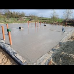 Viktoriia Nikolova Auto-LuX - Świeżo wylany fundament z wystającymi rurami instalacyjnymi i pionowymi oranżowymi rurami, zabezpieczony folią na obrzeżach, w tle zielona trawa i drzewa.