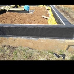 Viktoriia Nikolova Auto-LuX - Wrocław: Izolacja pionowa fundamentów z użyciem czarnej membrany hydroizolacyjnej, widoczna wykopana ziemia i przygotowany teren pod budowę.