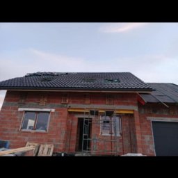 Viktoriia Nikolova Auto-LuX - Surowy budynek z czerwonej cegły z zamontowanymi oknami i dachem z czarnej dachówki, widoczny fragment rusztowania przy wejściu.