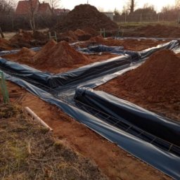 Viktoriia Nikolova Auto-LuX - Przygotowanie wykopu pod fundamenty domu z ułożoną folią hydroizolacyjną i zbrojeniem, w tle hałdy ziemi i zabudowa jednorodzinna.