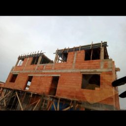 Viktoriia Nikolova Auto-LuX - Wznoszenie dwupiętrowego domu z czerwonej cegły, widoczne otwory okienne i drzwiowe, drewniane rusztowania i szalunki na górze, pochmurne niebo w tle.