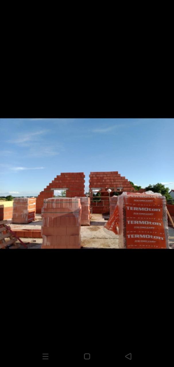 Wznoszone ściany budynku z czerwonej cegły, częściowo z otworami okiennymi, z paletami cegieł marki Termoton na pierwszym planie, błękitne niebo w tle.
