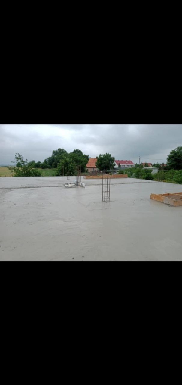 Wylany, świeży beton na płycie fundamentowej z wystającymi prętami zbrojeniowymi i drewnianym szalunkiem, widok na okoliczne drzewa i domy w oddali.