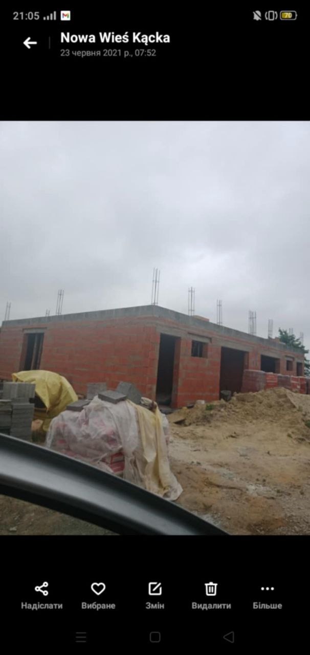 Wznoszona konstrukcja budynku z czerwonej cegły, widoczne zbrojenia wystające z dachu, materiały budowlane zabezpieczone folią na placu budowy, pochmurne niebo.