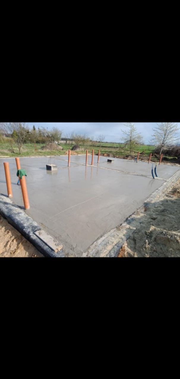 Świeżo wylany fundament z wystającymi rurami instalacyjnymi i pionowymi oranżowymi rurami, zabezpieczony folią na obrzeżach, w tle zielona trawa i drzewa.