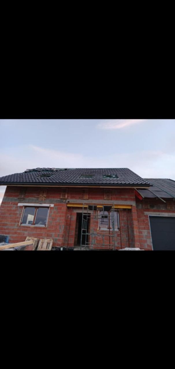 Surowy budynek z czerwonej cegły z zamontowanymi oknami i dachem z czarnej dachówki, widoczny fragment rusztowania przy wejściu.