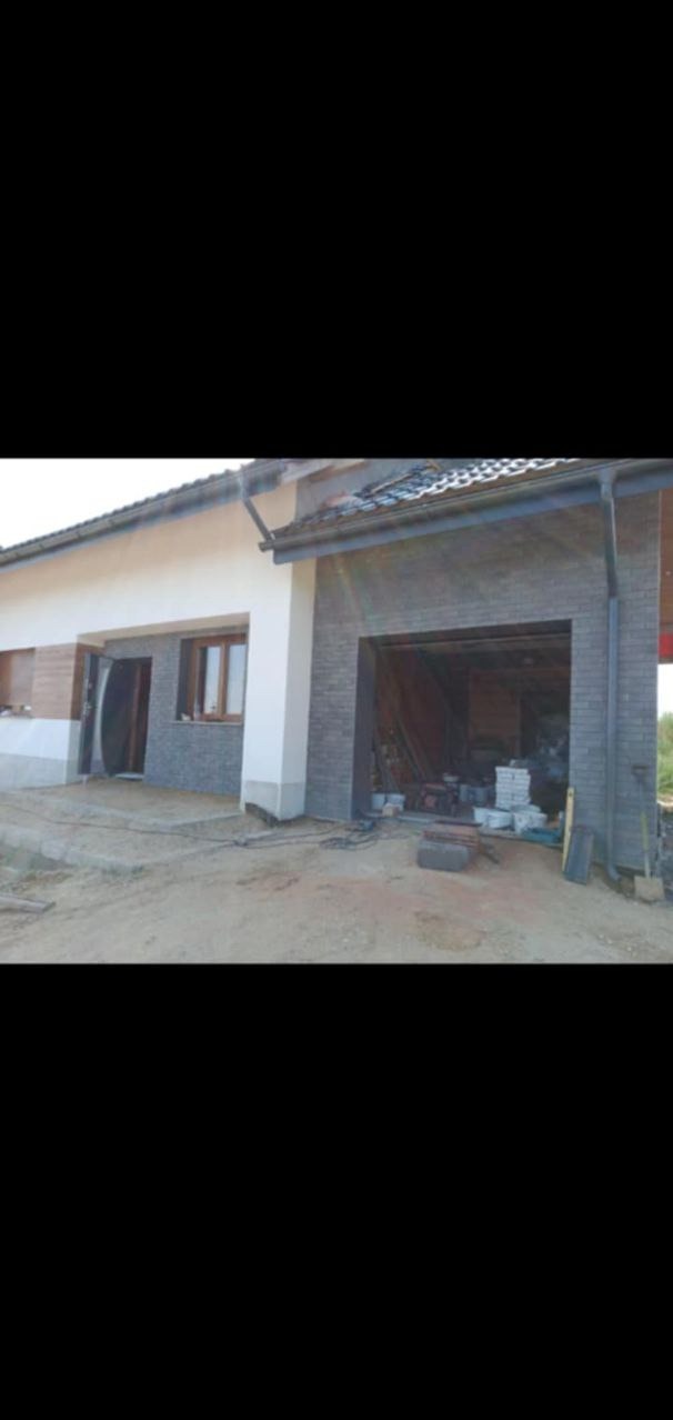 Niedokończony budynek z garażem w trakcie budowy, widoczne materiały budowlane i narzędzia w garażu, surowy teren wokół budynku.