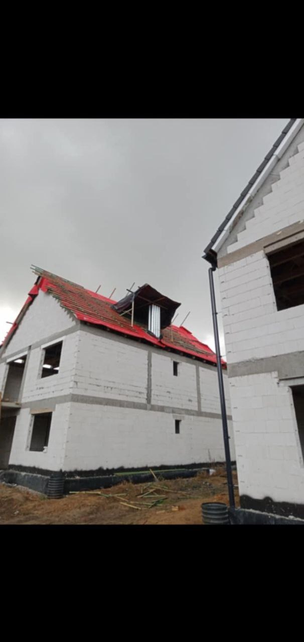 Wznoszenie ścian z bloczków betonu komórkowego na dwóch budynkach w stanie surowym otwartym, z widocznym deskowaniem dachu pokrytym czerwoną papą i kominem wentylacyjnym na jednym z nich, na tle...