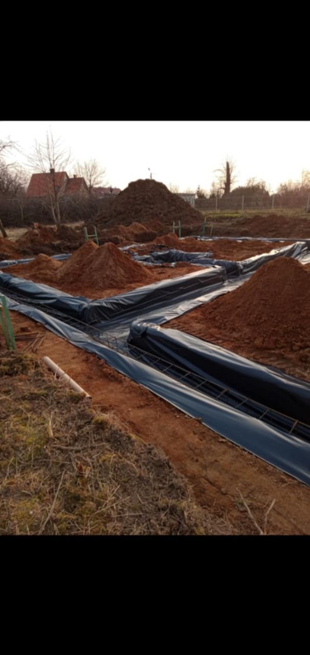 Przygotowanie wykopu pod fundamenty domu z ułożoną folią hydroizolacyjną i zbrojeniem, w tle hałdy ziemi i zabudowa jednorodzinna.