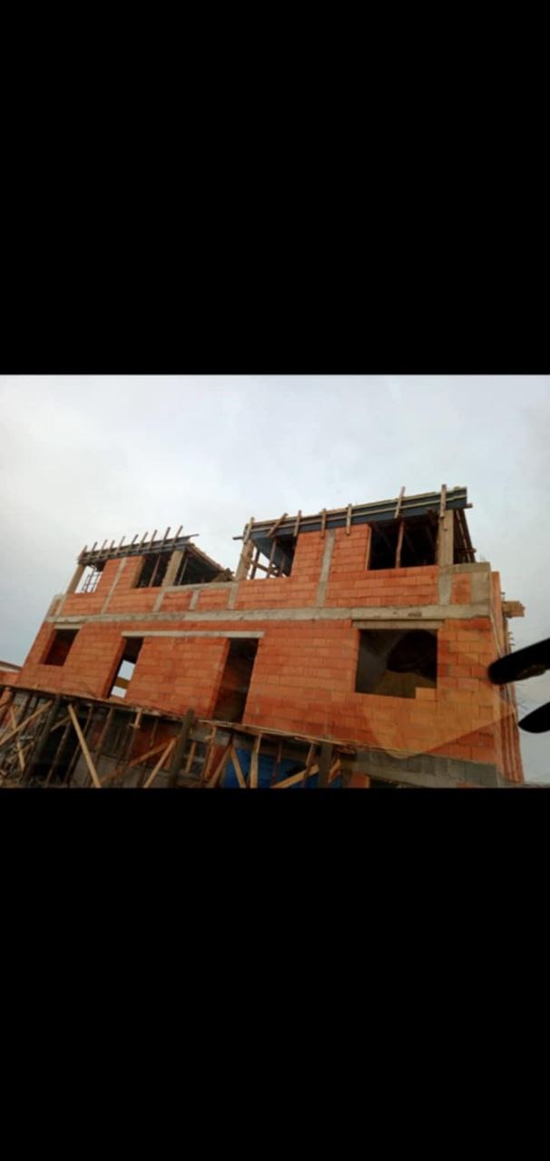 Wznoszenie dwupiętrowego domu z czerwonej cegły, widoczne otwory okienne i drzwiowe, drewniane rusztowania i szalunki na górze, pochmurne niebo w tle.