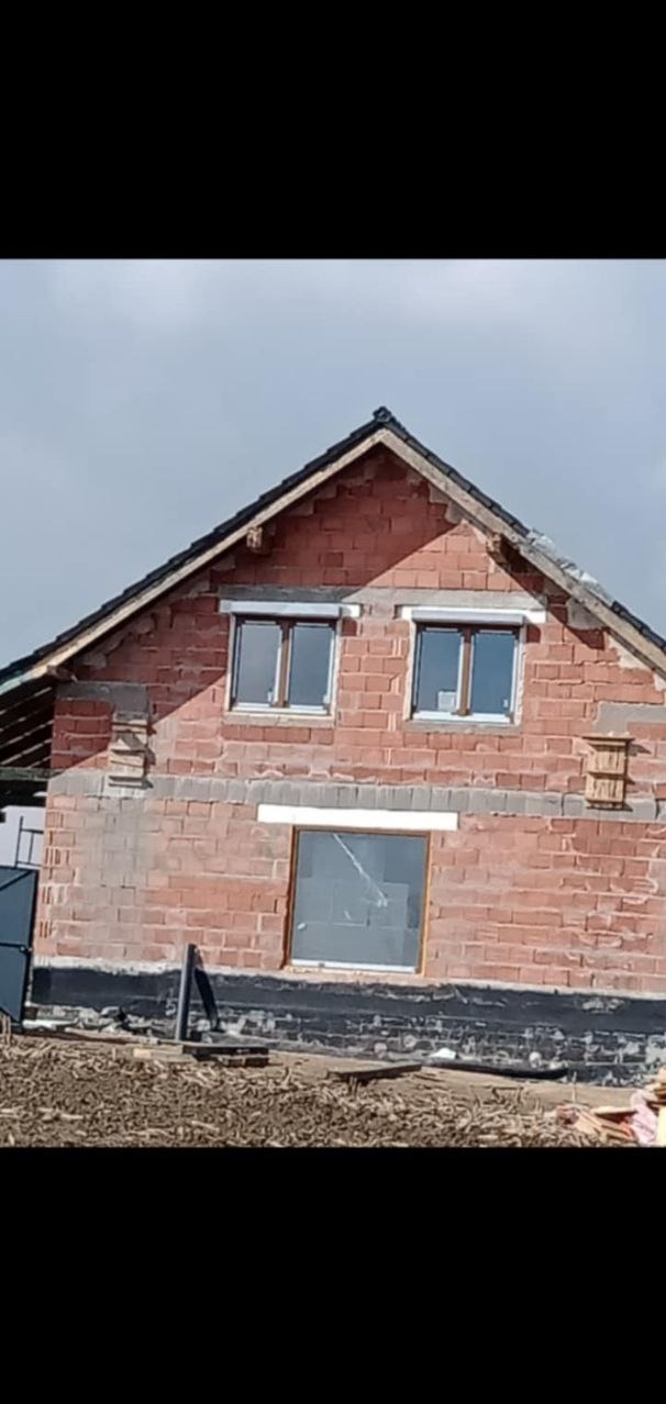 Wznoszony budynek z czerwonej cegły z zamontowanymi oknami i przygotowanym dachem, widoczne zabezpieczenie fundamentów czarną folią.