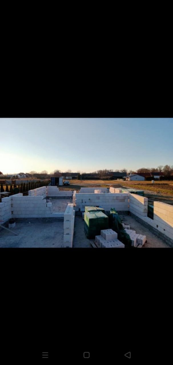 Wznoszone ściany parteru z bloczków betonu komórkowego na fundamencie, widoczne zapasy materiału budowlanego na placu budowy domu jednorodzinnego w słoneczny dzień.