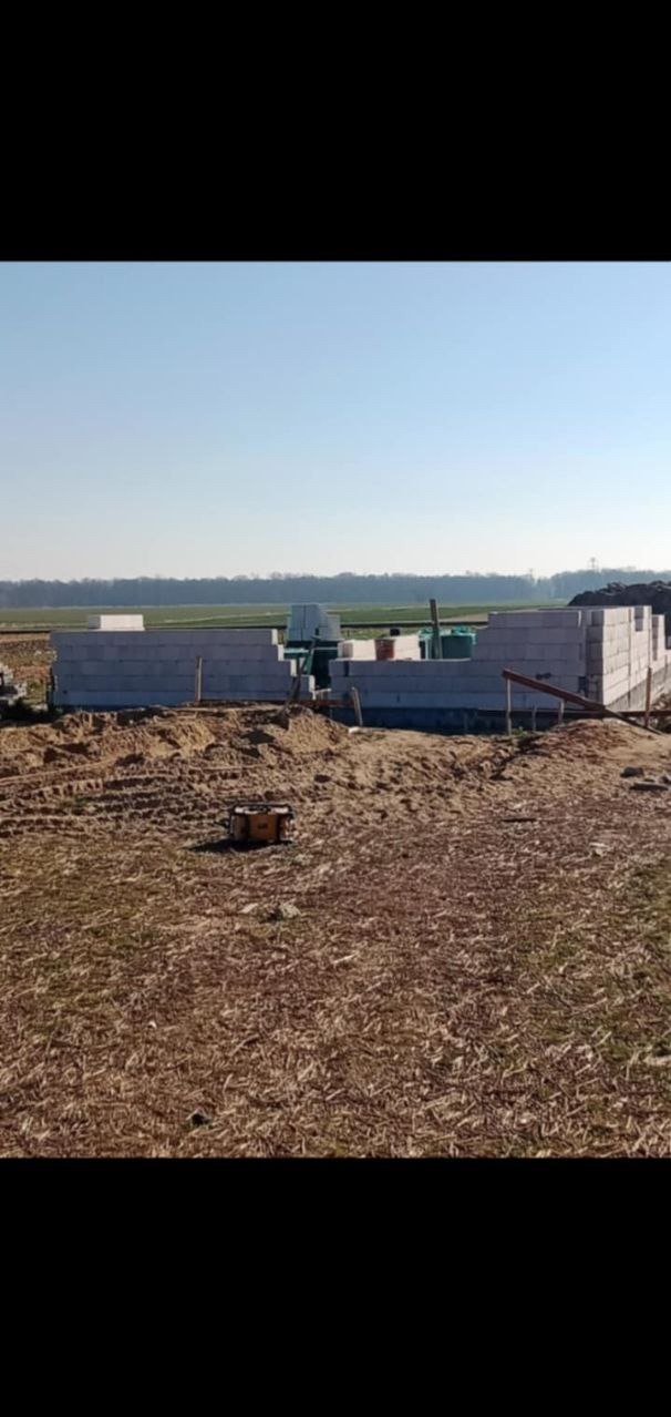 Wznoszone mury parterowego budynku z betonu komórkowego na tle pola i błękitnego nieba, widoczne narzędzia budowlane i żółta skrzynka narzędziowa.