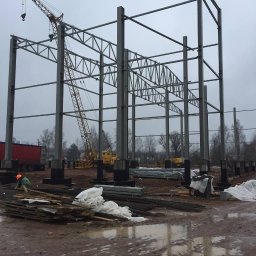 Roof Craft VASYL ANDROSHULIK - Konstrukcja stalowa hali w trakcie montażu, widoczne pionowe słupy, kratownice, żuraw budowlany, materiały budowlane składowane na błotnistym terenie, pracownik w kasku.