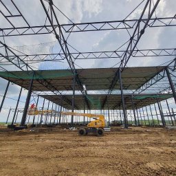 Roof Craft VASYL ANDROSHULIK - Konstrukcja stalowa hali w Wieliczce w trakcie montażu, widoczny podnośnik koszowy z pracownikiem w pomarańczowej koszulce, zabezpieczenia siatką oraz częściowo zamontowane płyty dachowe.
