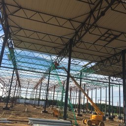 Roof Craft VASYL ANDROSHULIK - Montaż dachu hali z płyt warstwowych z użyciem podnośnika koszowego, widoczne stalowe elementy konstrukcyjne, zabezpieczenia siatką i stos płyt na ziemi.