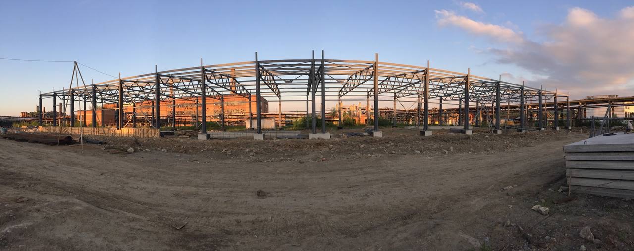 Szeroki kadr na konstrukcję stalową hali w trakcie budowy, widoczne fundamenty i elementy szkieletu, w tle budynki przemysłowe i niebo o zachodzie słońca.
