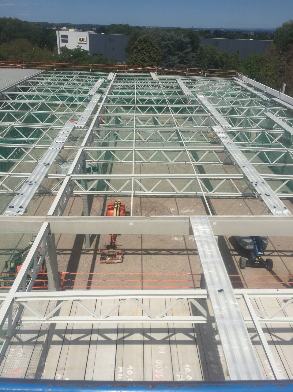 Widok z góry na konstrukcję stalową dachu hali w trakcie montażu, z widocznymi podnośnikami koszowymi i zabezpieczeniem siatką, w tle budynek z napisem MODEL.