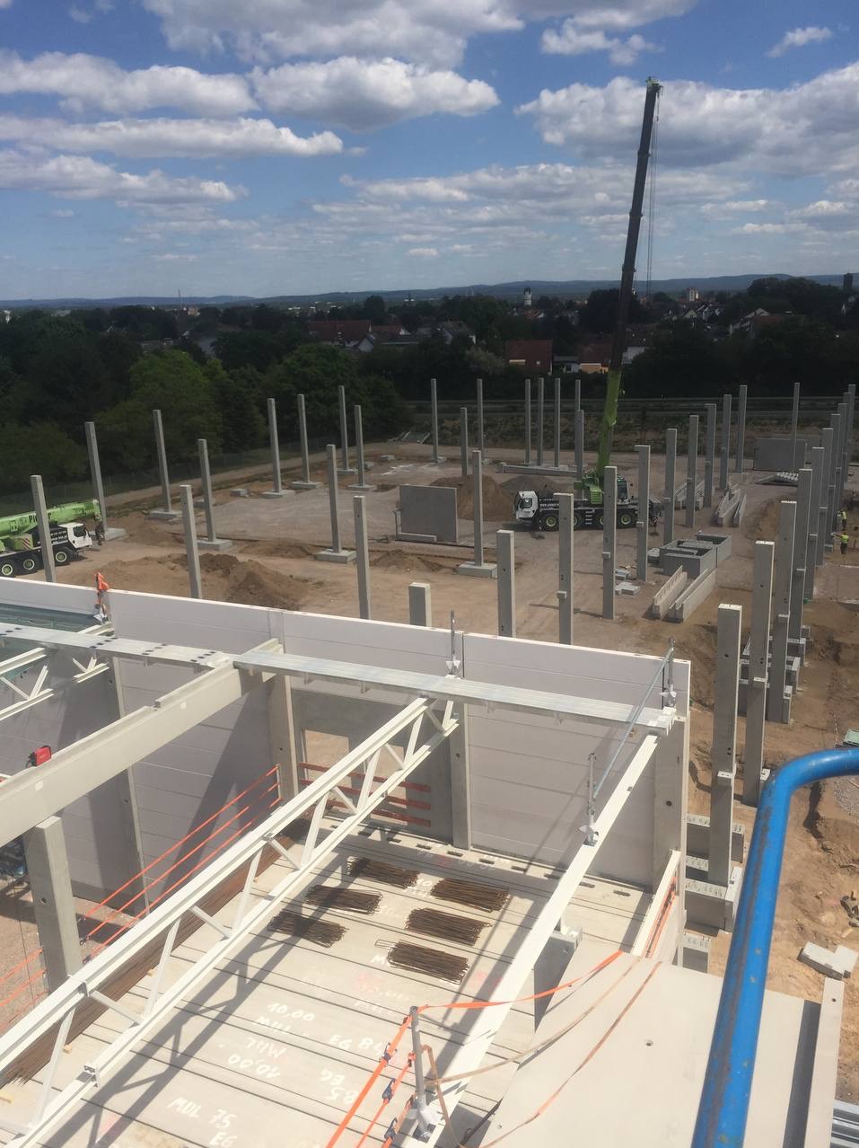 Budowa hali z betonu: widok z góry na konstrukcję szkieletową z kolumnami i ścianami, w tle żuraw budowlany i ciężarówki z betonem.