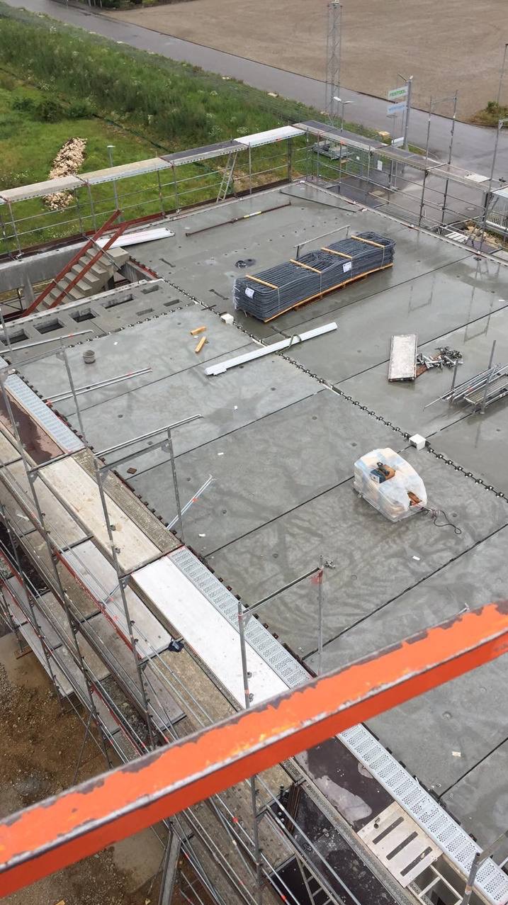 Widok z góry na mokry dach w trakcie budowy, z widocznymi rusztowaniami, ułożonymi płytami, zwojami stali zbrojeniowej i narzędziami.