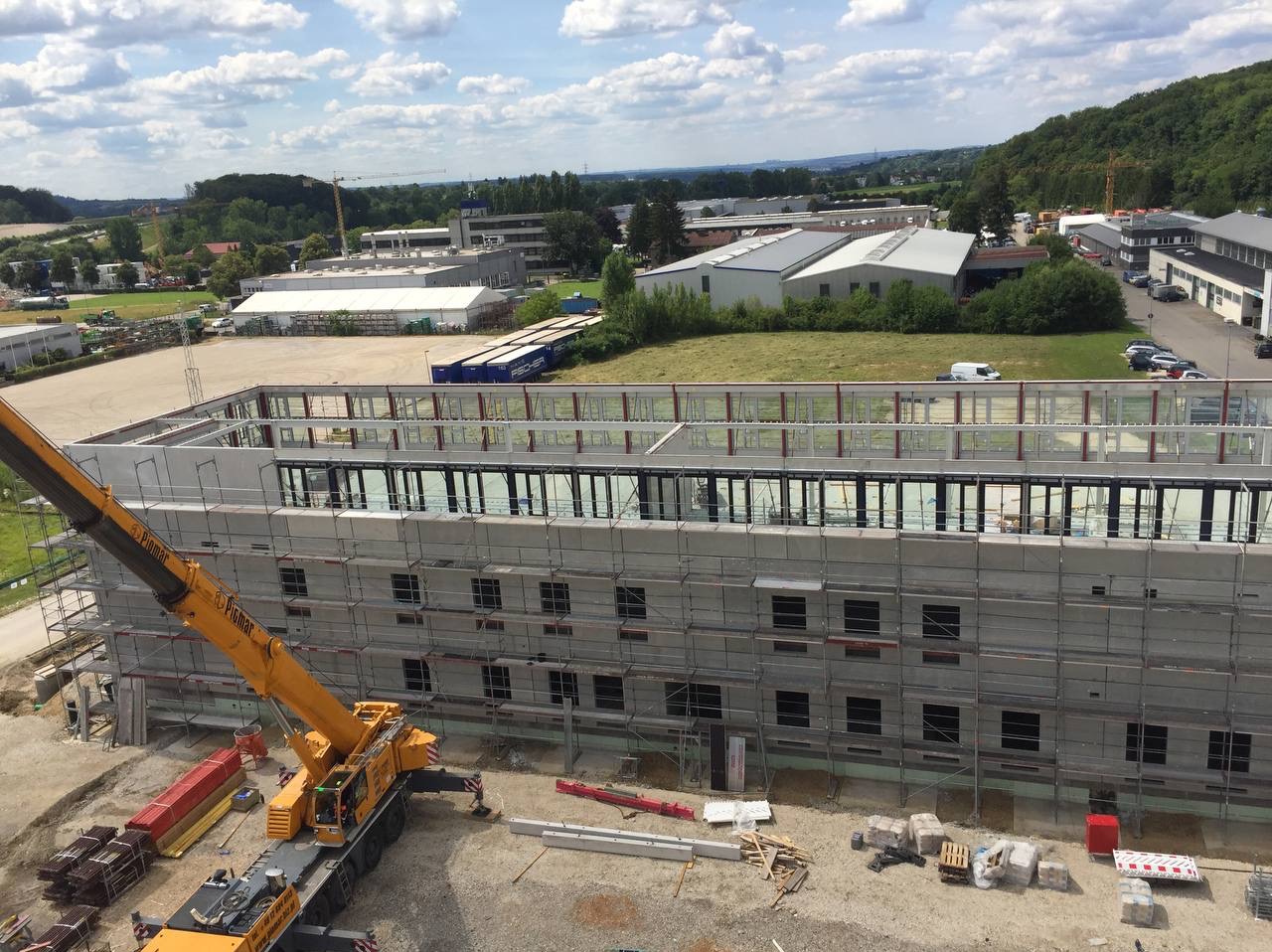 Budowa hali z widocznym rusztowaniem, dźwigiem, elementami konstrukcyjnymi i otaczającym terenem przemysłowym w słoneczny dzień.