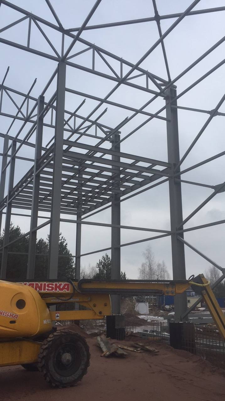 Wznoszenie konstrukcji stalowej hali, widoczny szkielet budynku z belek i słupów, platforma robocza podnośnika koszowego w kolorze żółtym, teren budowy w tle.