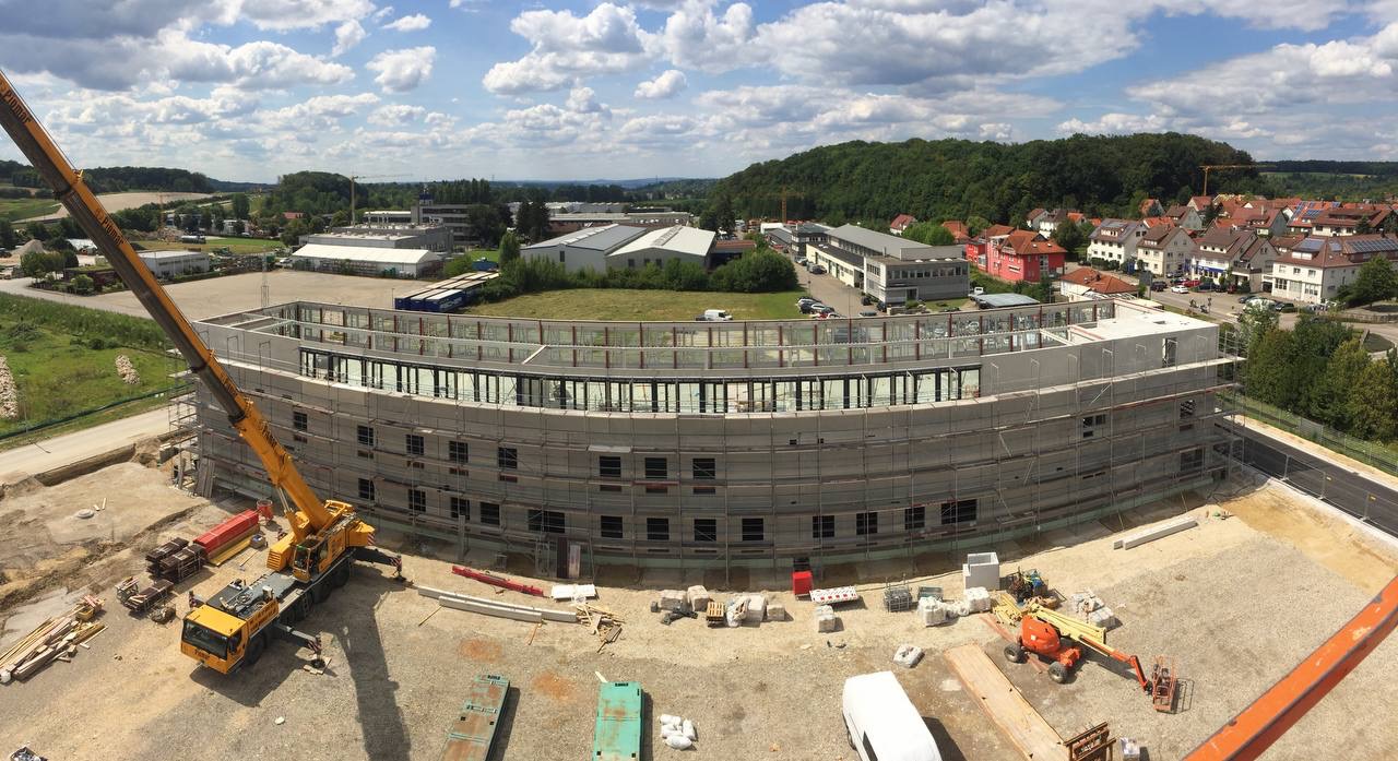 Budowa budynku o zaokrąglonym kształcie z użyciem żurawia, widoczne rusztowania i materiały budowlane na placu budowy, w tle zabudowa miejska i zalesione wzgórza.