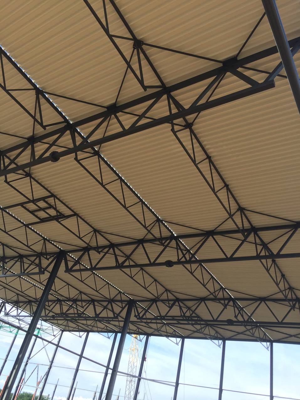 Wnętrze hali w trakcie budowy, widok na stalową konstrukcję dachu z beżową blachą trapezową, w tle widoczny dźwig budowlany i błękitne niebo.