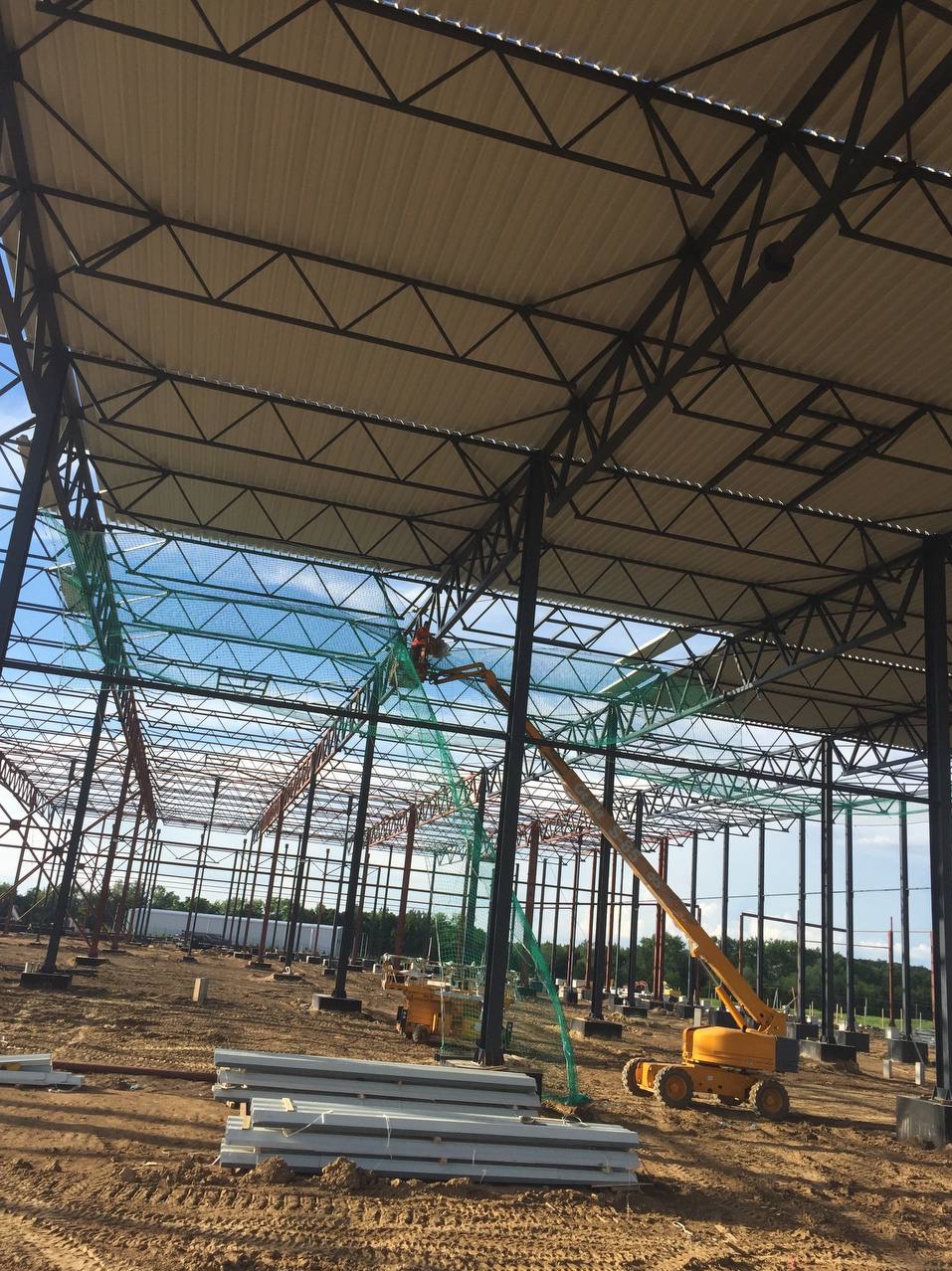 Montaż dachu hali z płyt warstwowych z użyciem podnośnika koszowego, widoczne stalowe elementy konstrukcyjne, zabezpieczenia siatką i stos płyt na ziemi.