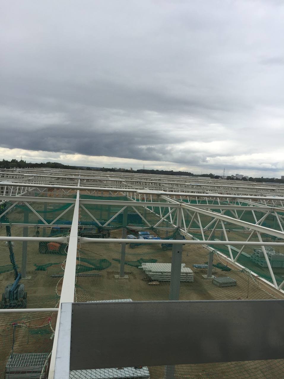 Konstrukcja stalowa hali w trakcie budowy, widoczne elementy szkieletu, zabezpieczenia siatką, materiały budowlane i pochmurne niebo w tle.