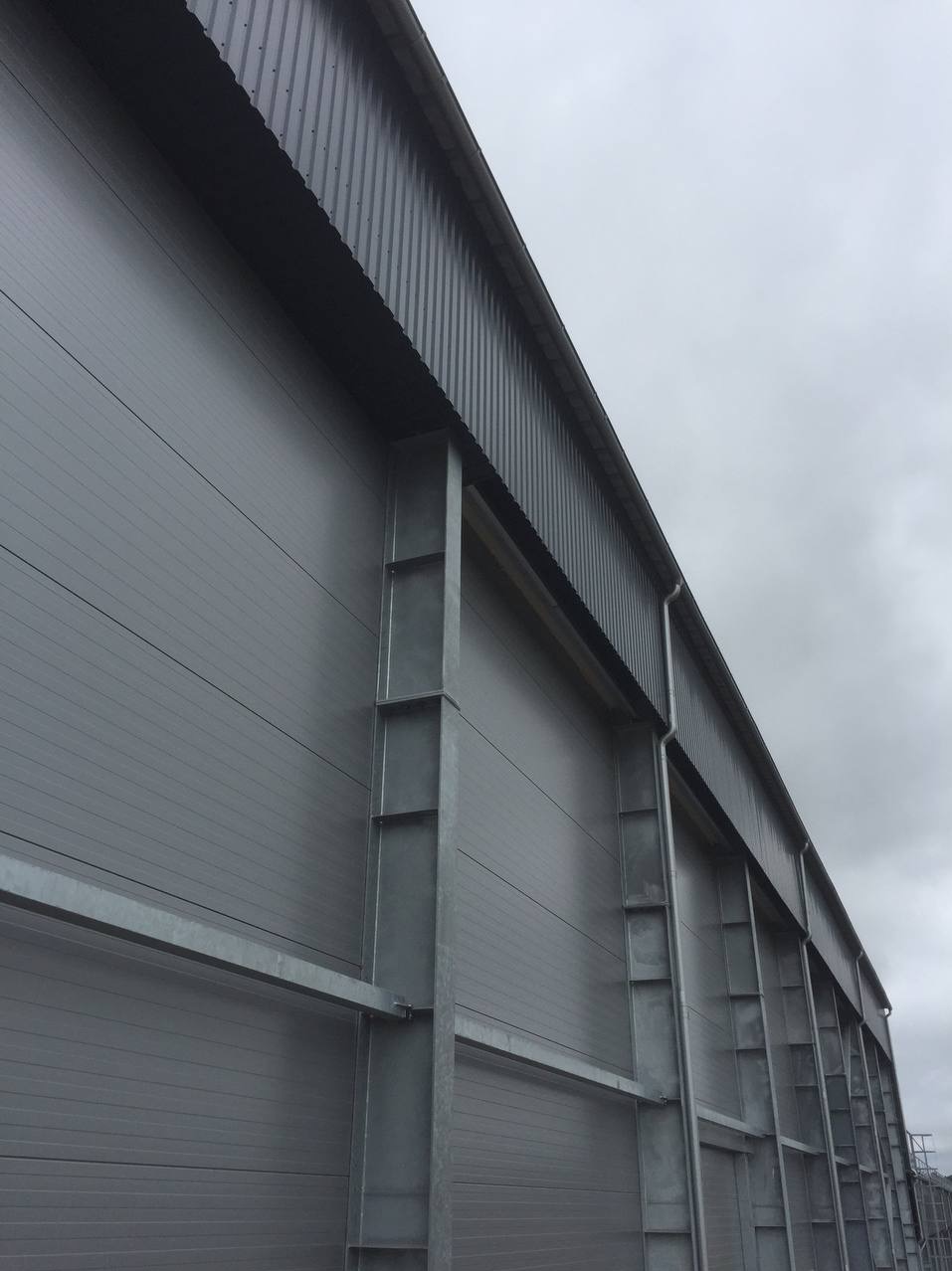 Szara hala przemysłowa z paneli warstwowych, widok z dołu na fragment ściany i dachu, elementy konstrukcyjne metalowe, pochmurne niebo w tle.