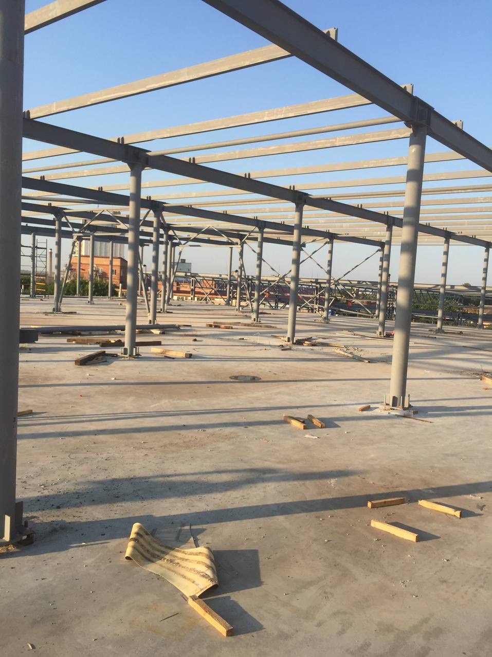 Surowa konstrukcja stalowa hali w trakcie budowy, widoczne szare słupy i poprzeczne belki, betonowa posadzka z resztkami materiałów budowlanych, w tle budynki i niebieskie niebo.