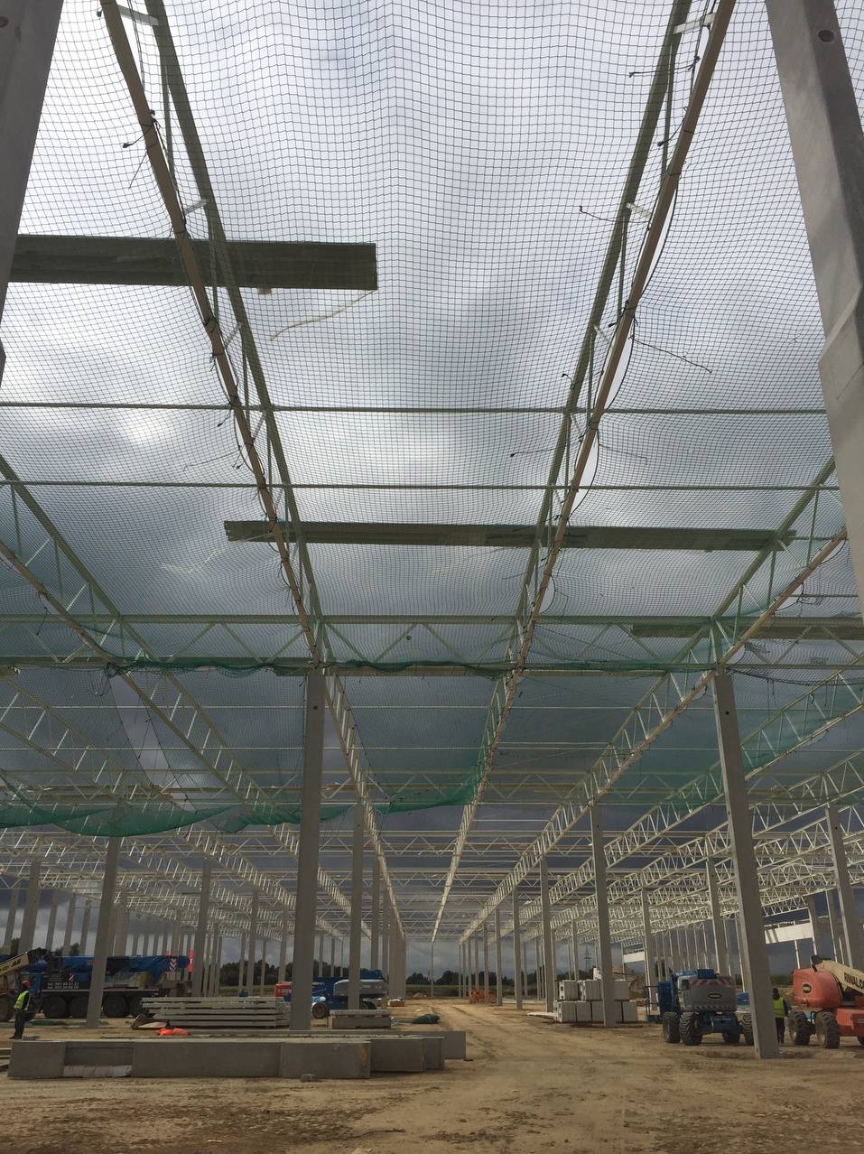 Wnętrze budowanej hali stalowej z widoczną konstrukcją nośną, betonowymi słupami i siatką zabezpieczającą pod dachem, maszyny budowlane i materiały na placu budowy.