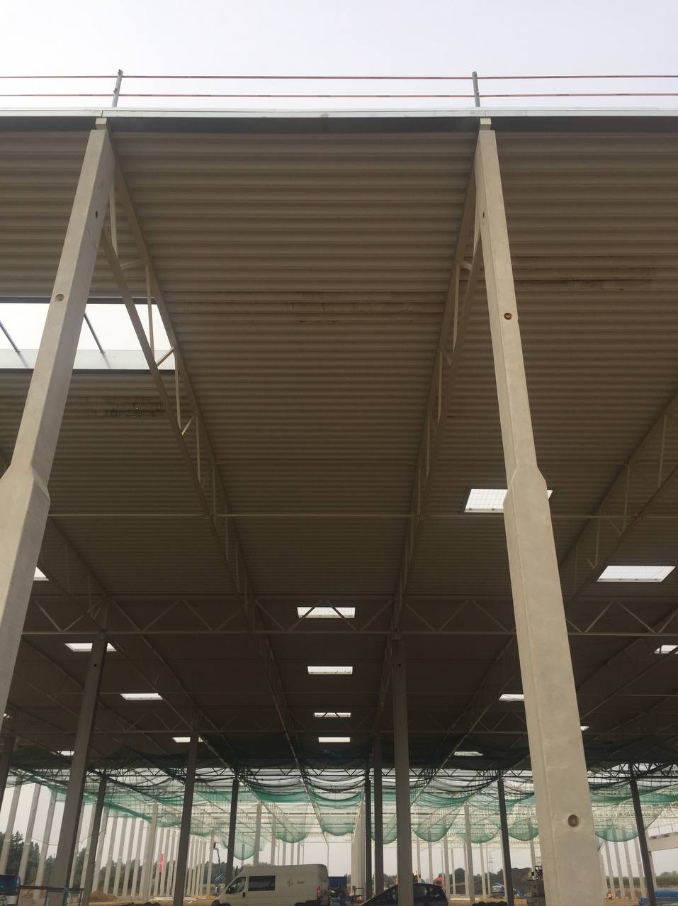 Wnętrze hali w trakcie budowy, widoczne stalowe konstrukcje, betonowe słupy i płyty warstwowe tworzące dach, zabezpieczenia siatką na dole.