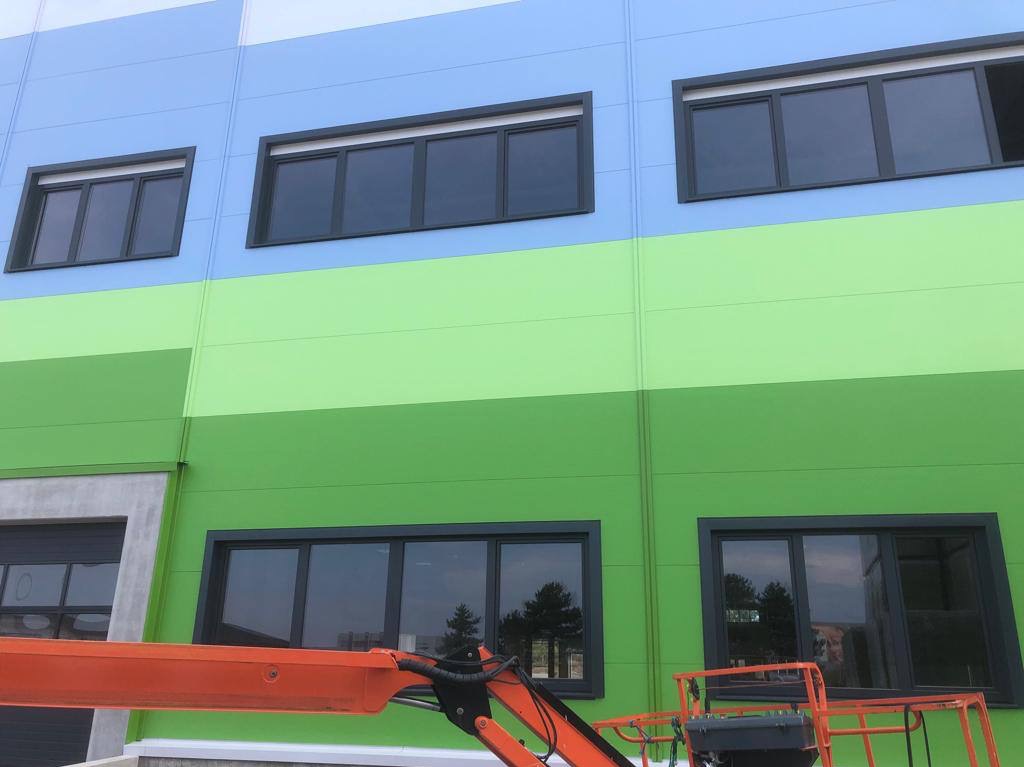 Fragment pomalowanej na niebiesko i zielono hali z zamontowanymi oknami, widziany z podnośnika koszowego.