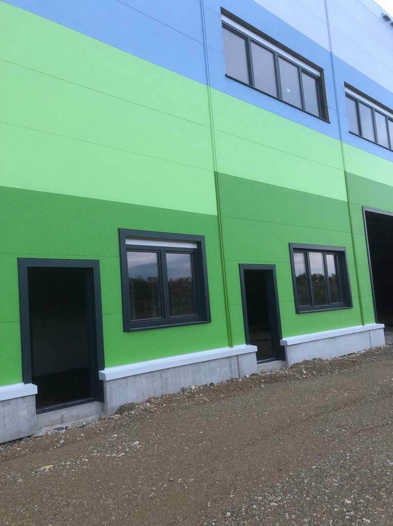 Nowo wybudowana hala z płyt warstwowych w odcieniach niebieskiego i zielonego, z widocznymi oknami i otworami drzwiowymi, teren wokół jeszcze niezagospodarowany.