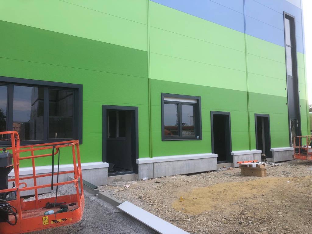 Budynek przemysłowy z zielono-niebieską elewacją z płyt warstwowych, widoczne okna w czarnych ramach, drzwi wejściowe i podnośnik koszowy w trakcie prac wykończeniowych.