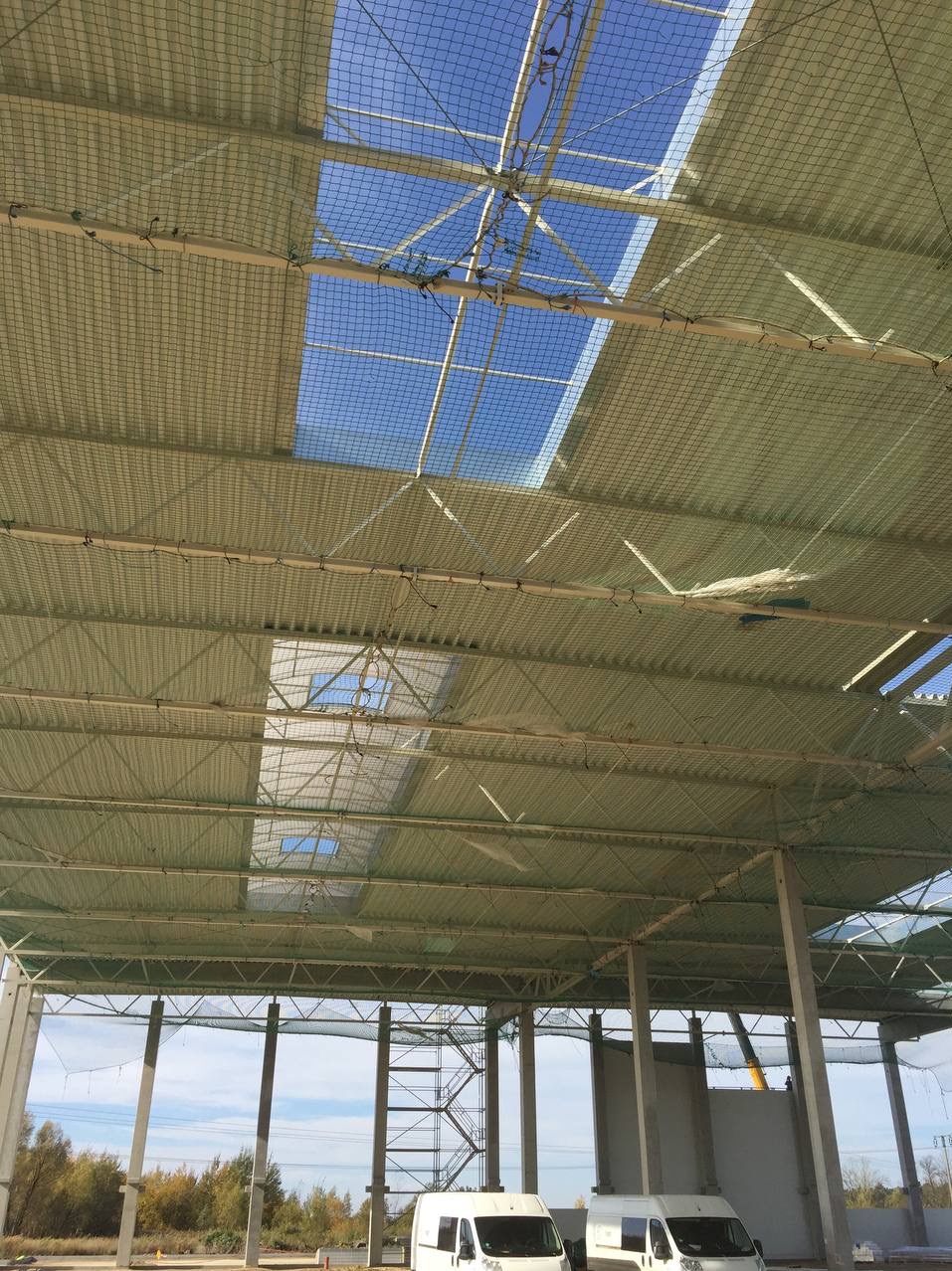Wnętrze hali przemysłowej w trakcie budowy z widoczną konstrukcją dachu, siatką zabezpieczającą i wpadającym światłem słonecznym przez otwory. Na zewnątrz zaparkowane białe busy.