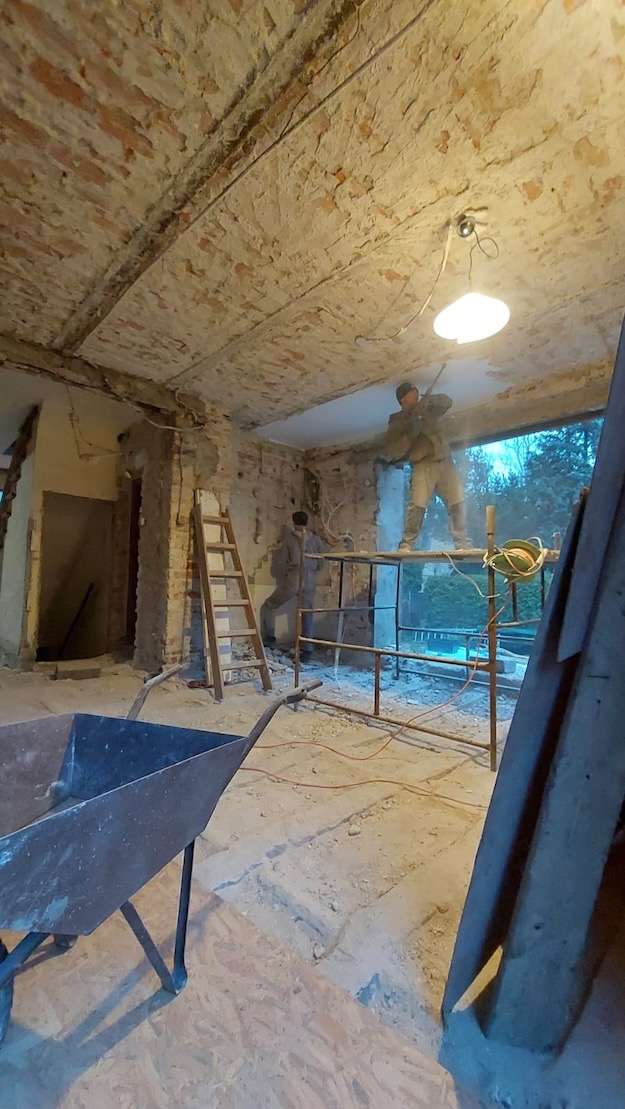 Wnętrze w trakcie prac remontowych: robotnicy na rusztowaniu, ceglany sufit z resztkami tynku, gruz na podłodze, taczka w lewym dolnym rogu, drabina oparta o ścianę.